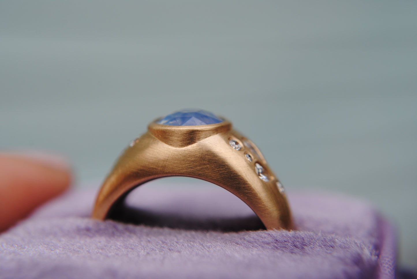 Ring with blue center stone and side diamonds against a blurred purple background shown on it's side