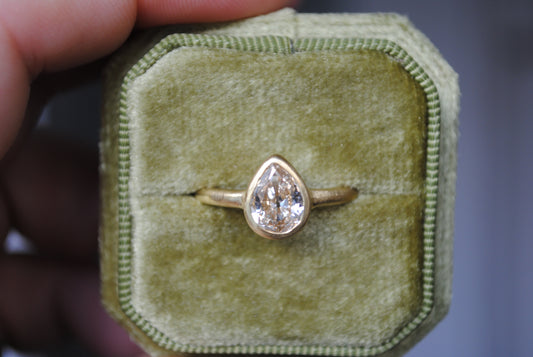 A champagne-toned, antique pear cut diamond ring in a matte finish, set in a bezel setting on a green velvet-lined box.