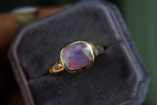 A pink opalescent sapphire ring with gold band, displayed on a gray cushion.