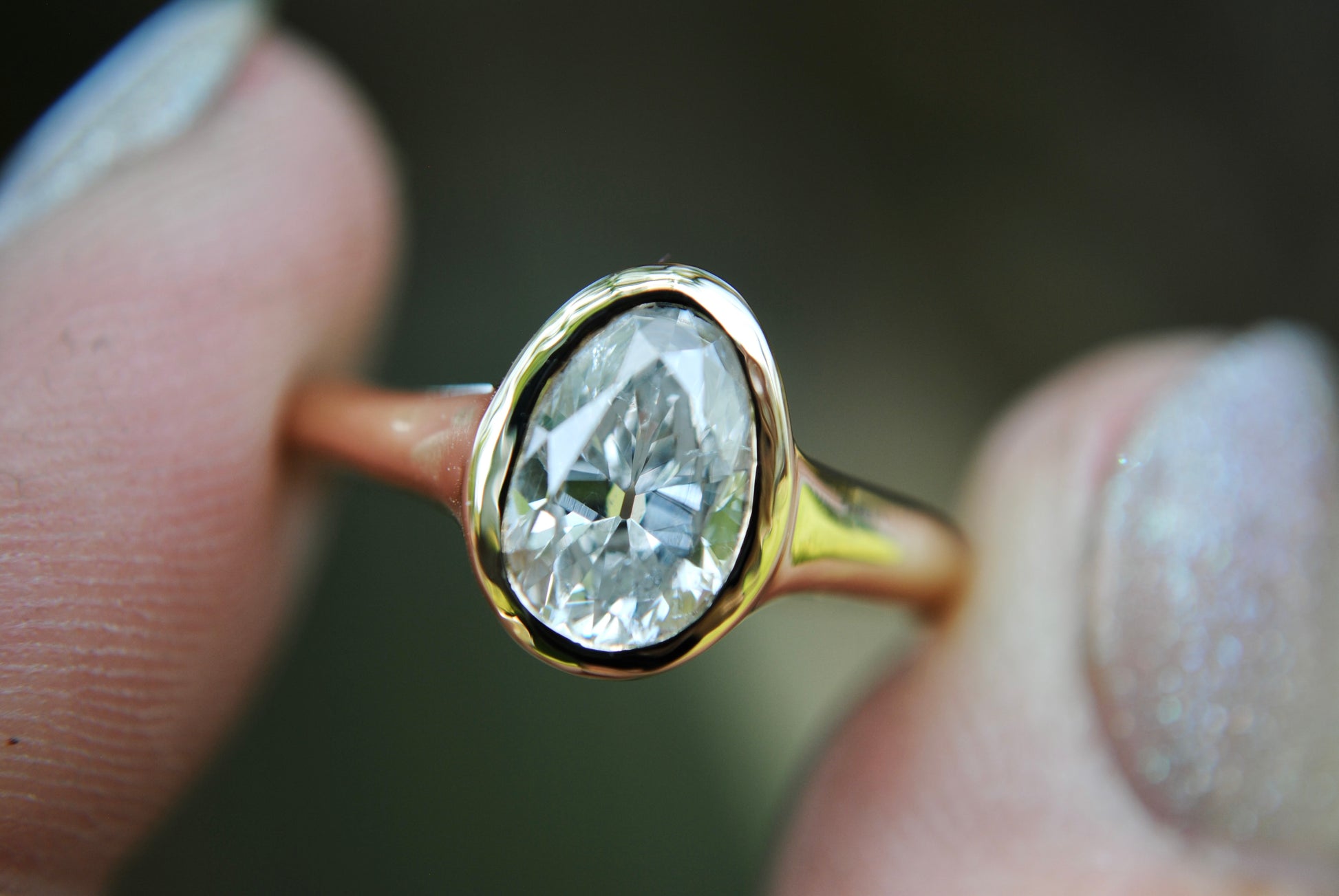 Gold ring with a large diamond held between two fingers against a blurred background