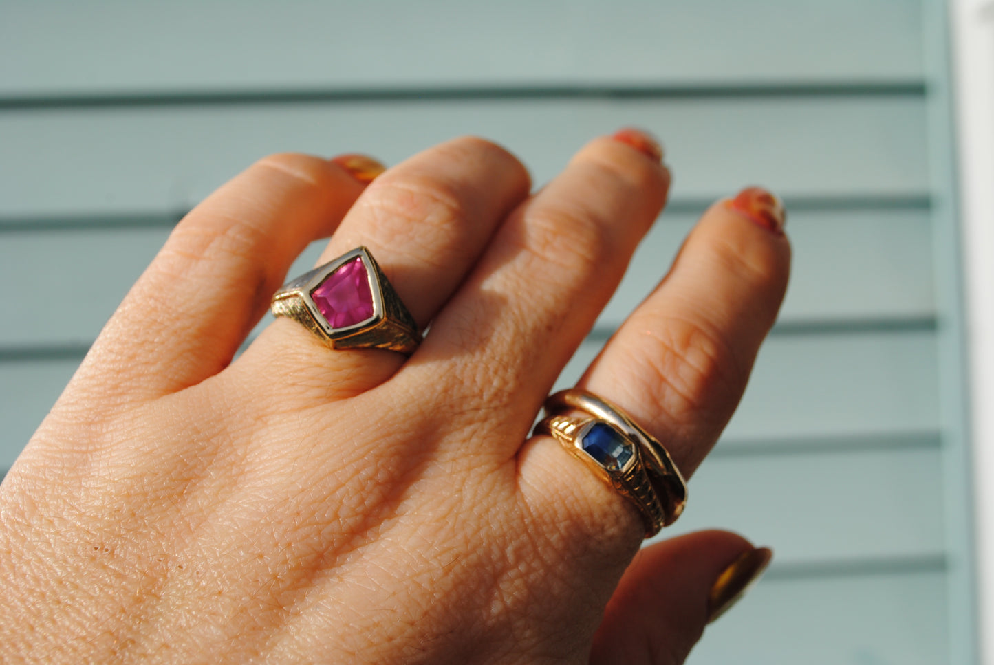 Hand wearing two gold rings with gemstones against a light blue background