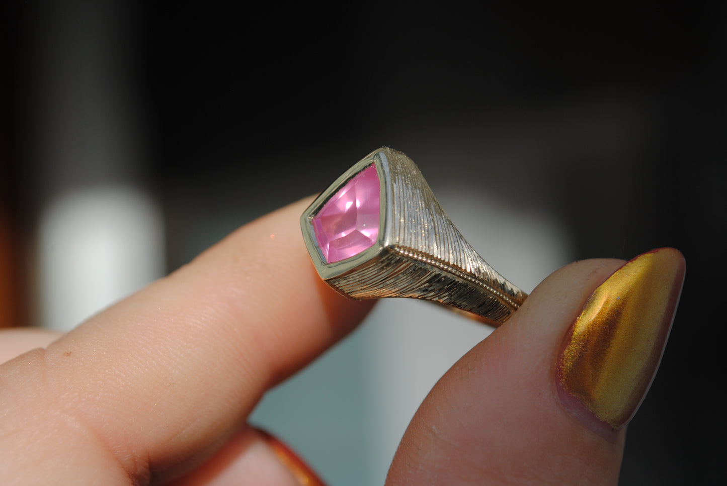 Gold ring with a pink gemstone held by a hand with gold nail polish.