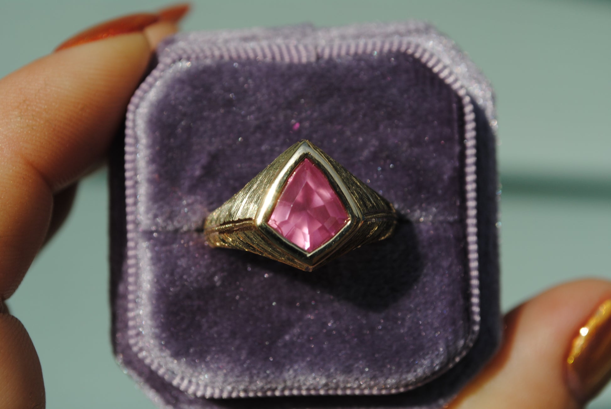 Gold ring with a pink gemstone on purple velvet in a hand.