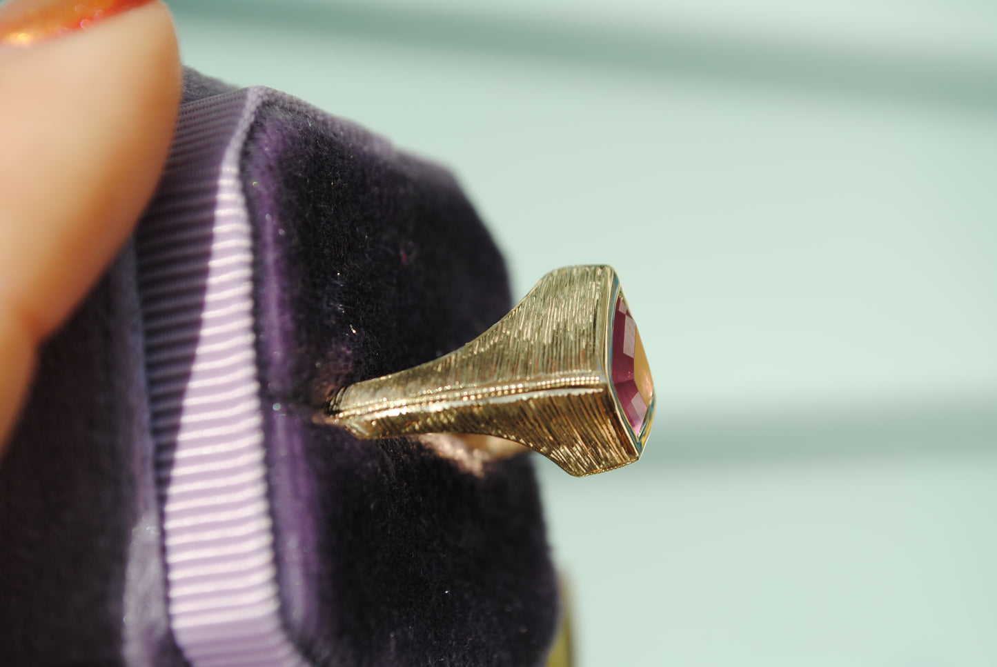 Gold ring with a red gemstone held by a hand on a purple cushion.