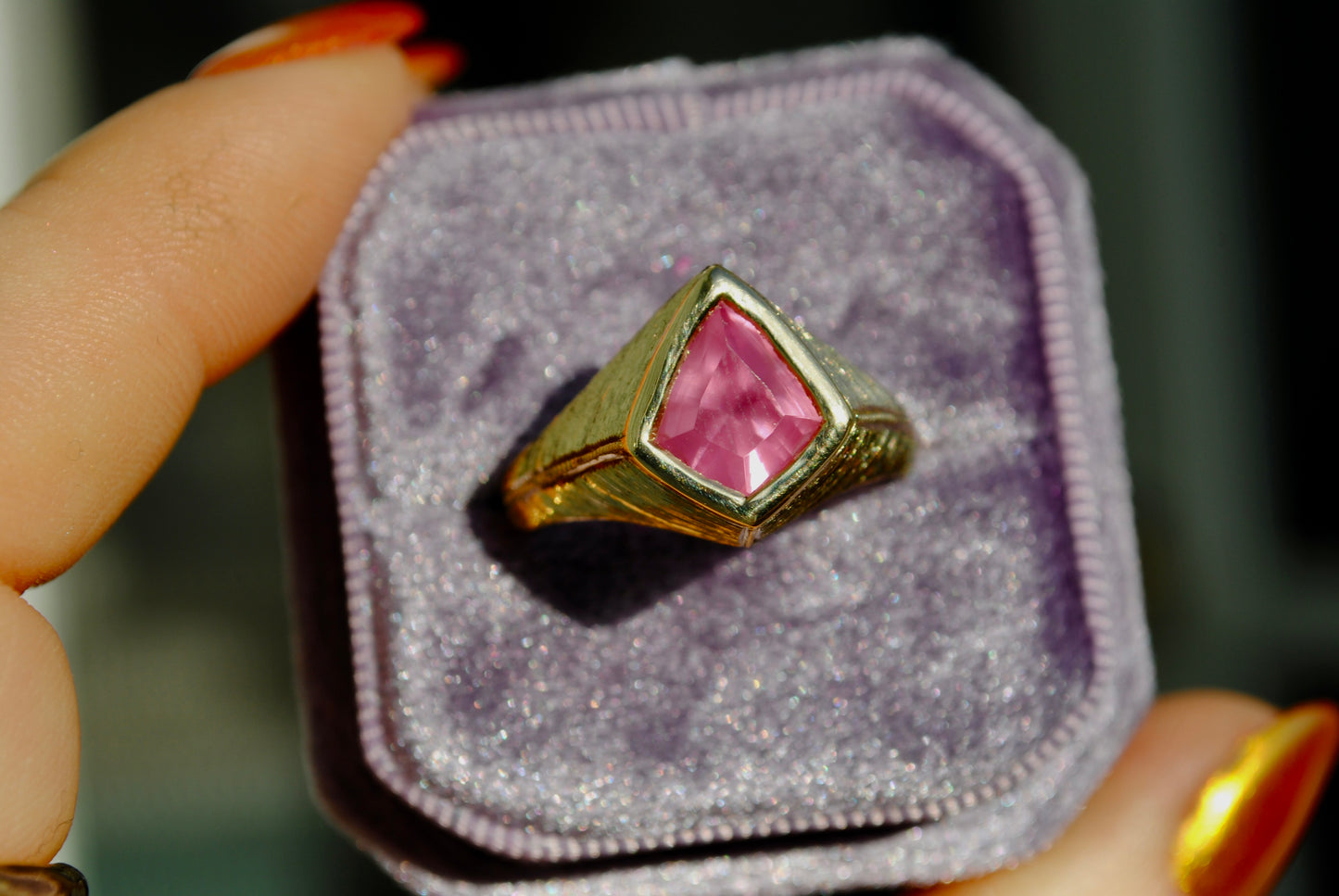 Gold ring with a pink gemstone in a purple velvet box held by a person with orange nail polish.