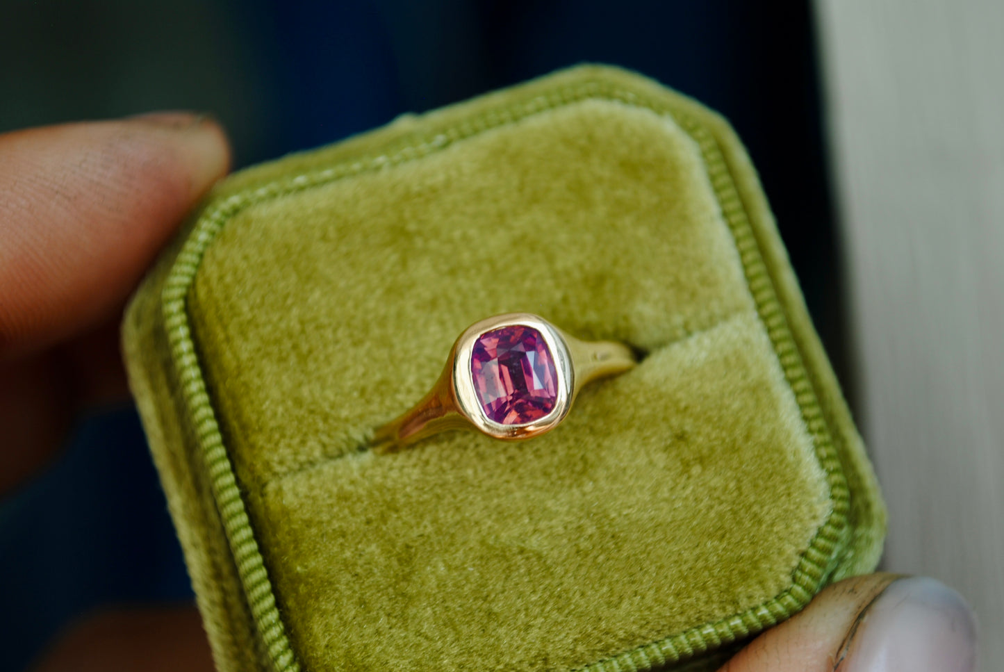 Gold ring with a pink gemstone in a green velvet box held by a hand.