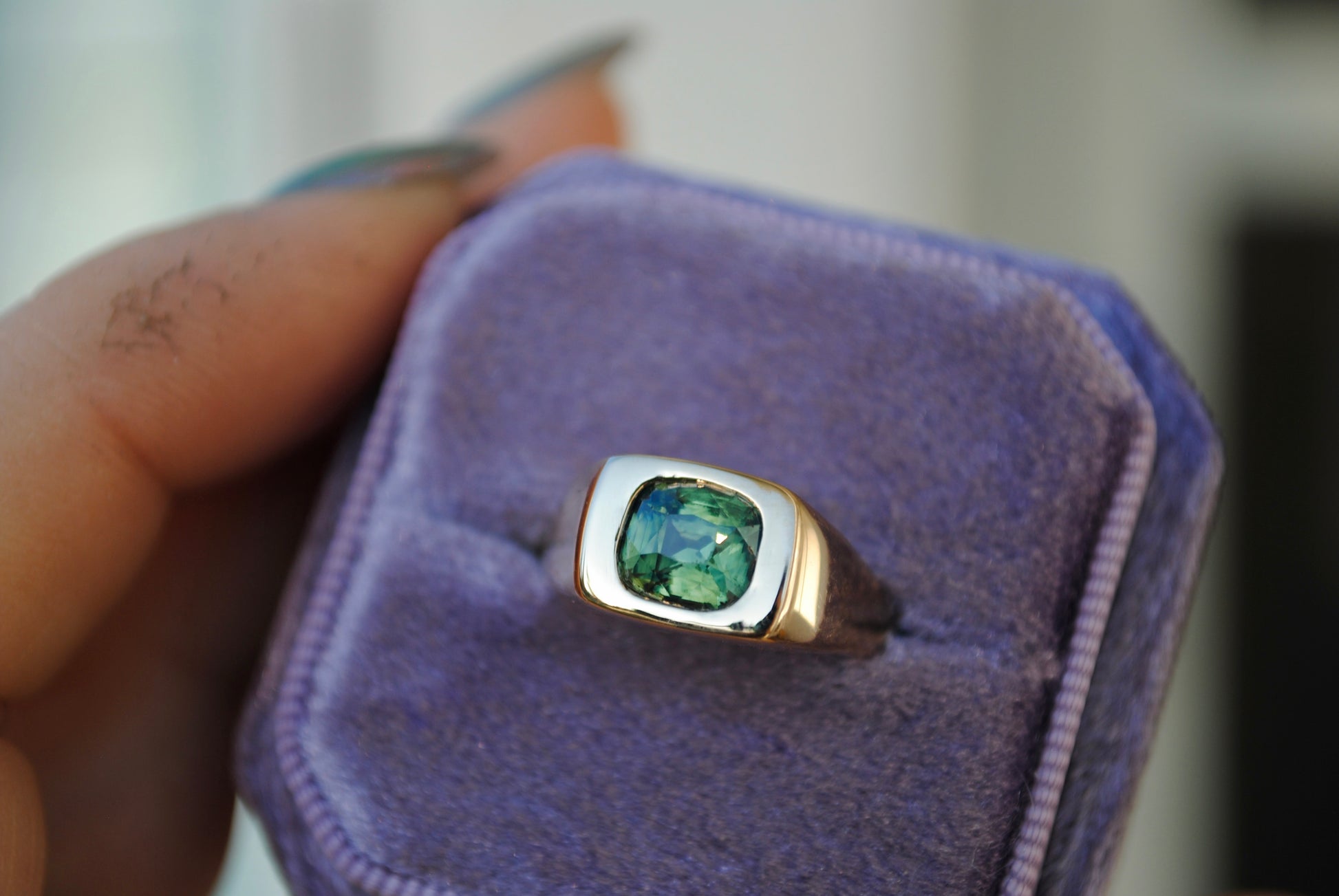 Gold ring with a green gemstone in a purple box held by a hand.
