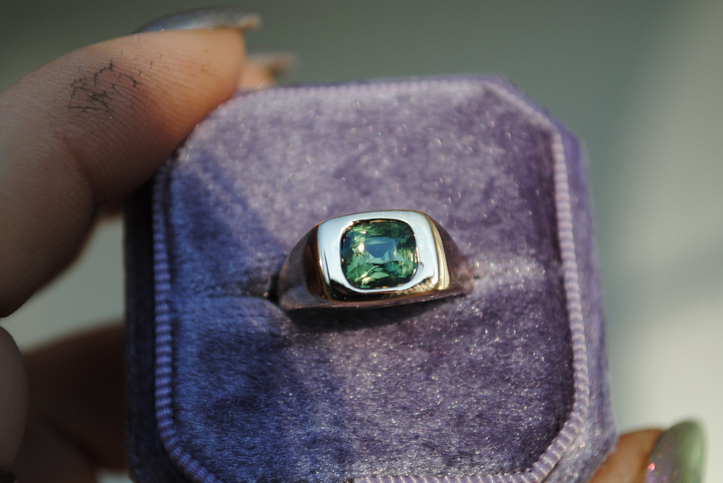 Ring with a green gemstone in a purple velvet box held by a hand.