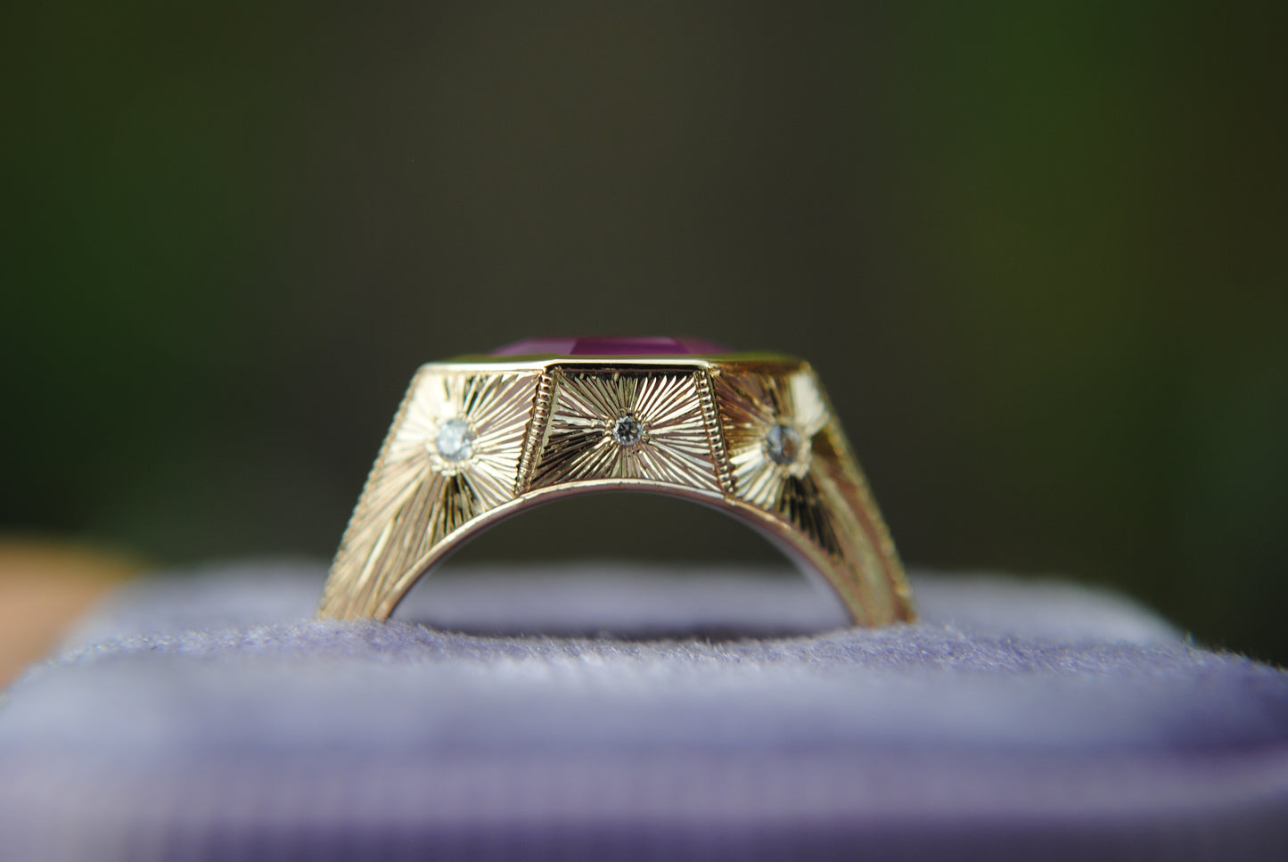 Gold ring with a purple stone on a purple cushion against a blurred green background
