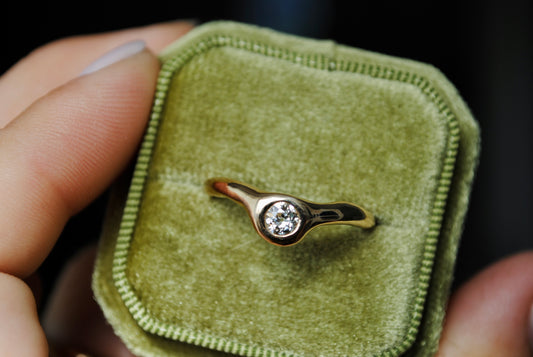 Gold ring with a diamond on a green velvet cushion held by a hand.