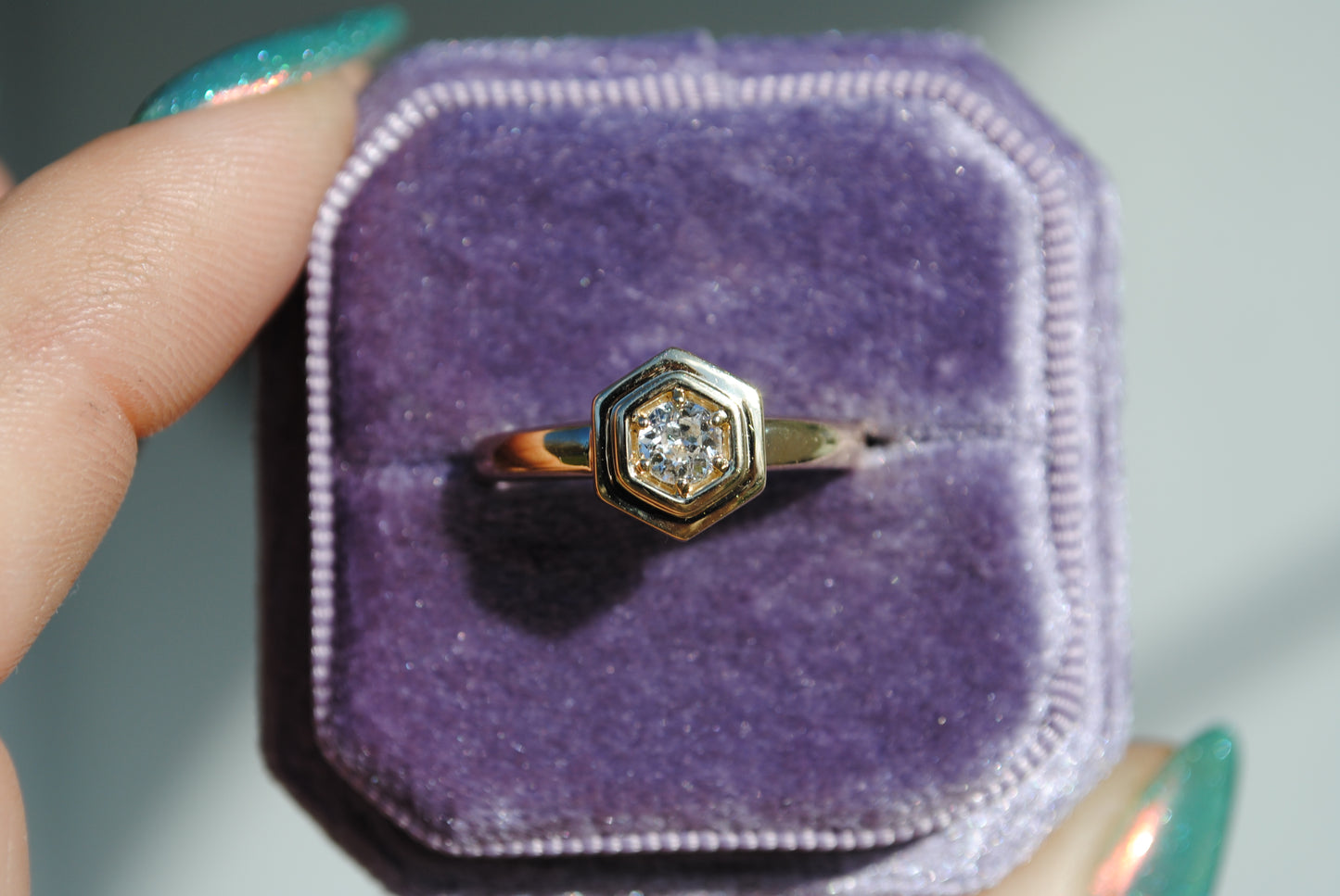 Gold ring with a diamond in a purple box held by a hand.