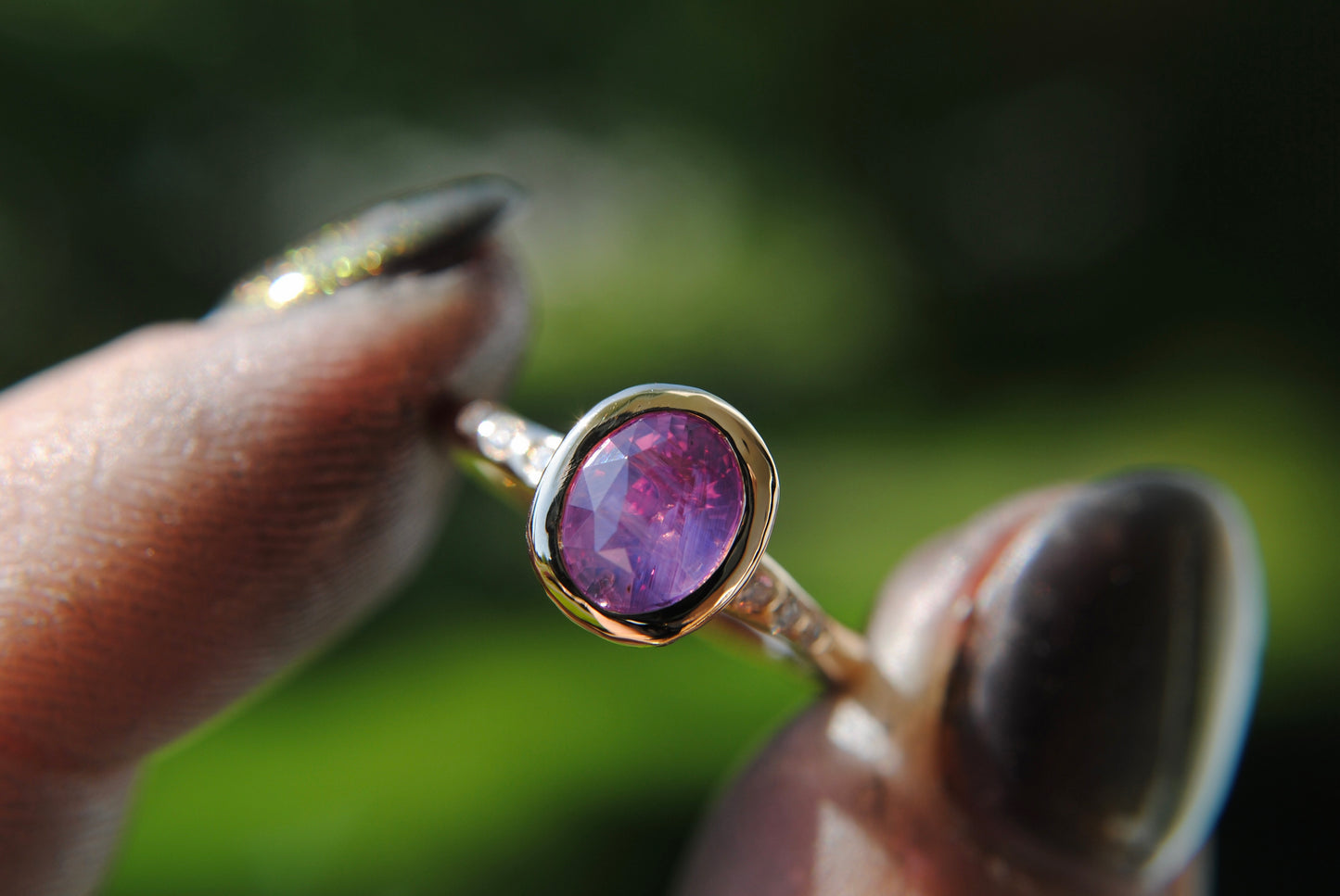 Gold ring with a purple gemstone held between fingers against a blurred green background