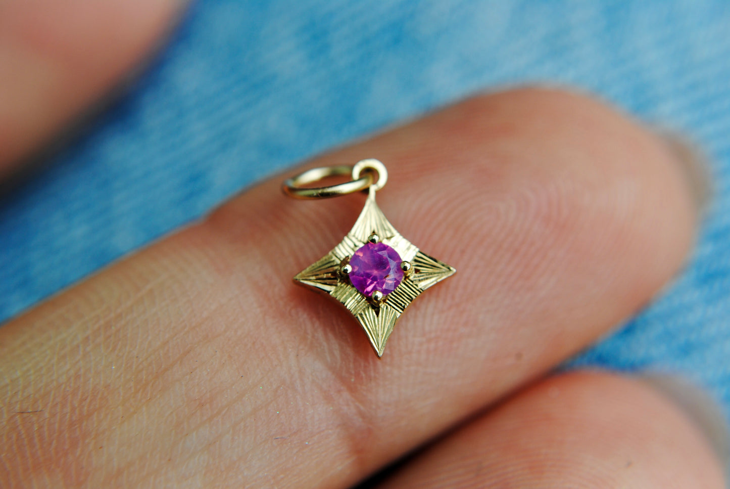Gold pendant with a purple gemstone held between fingers against a blue fabric background
