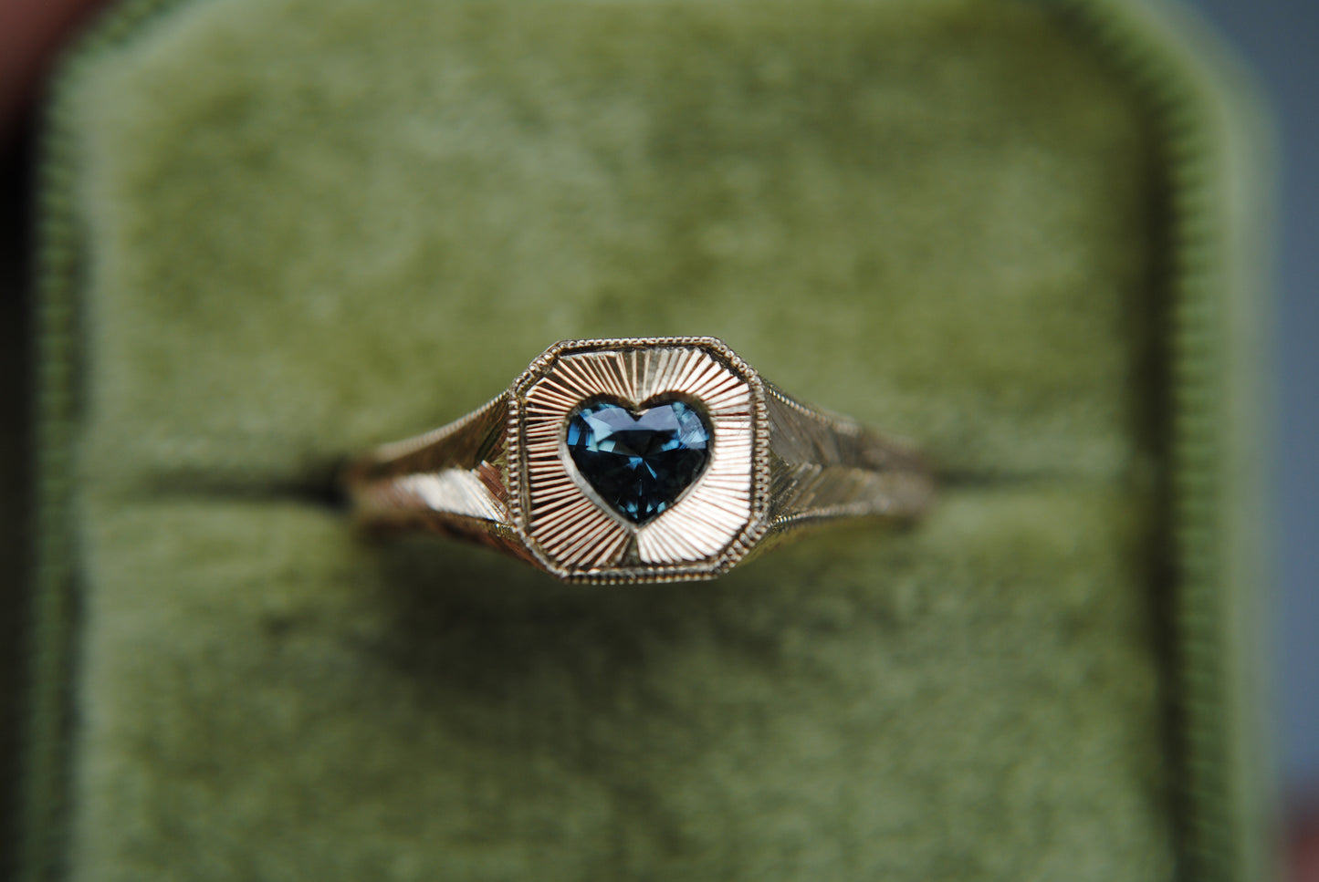 Gold ring with a heart-shaped blue gemstone on a green cushioned background