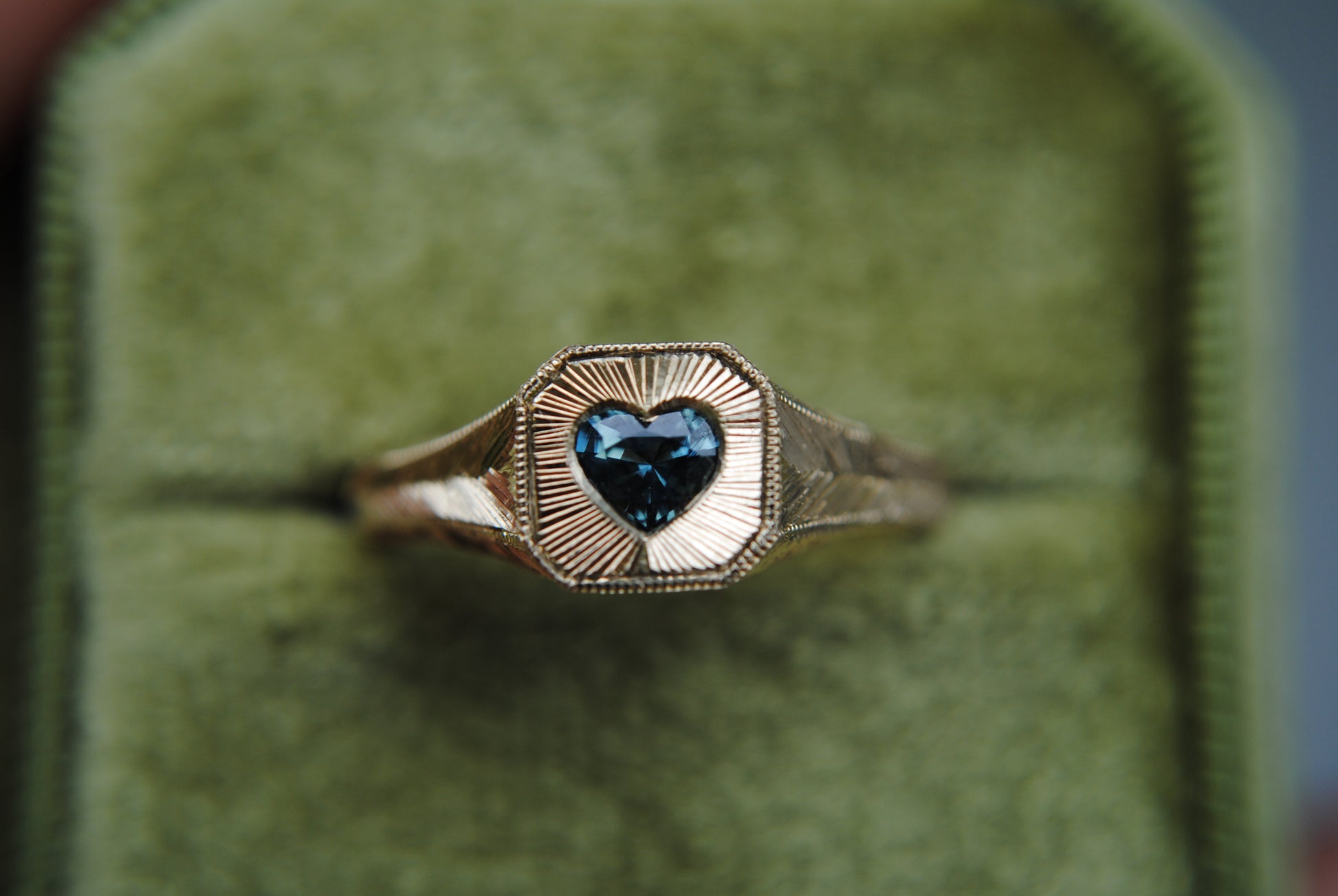 Gold ring with a heart-shaped blue gemstone on a green cushioned background