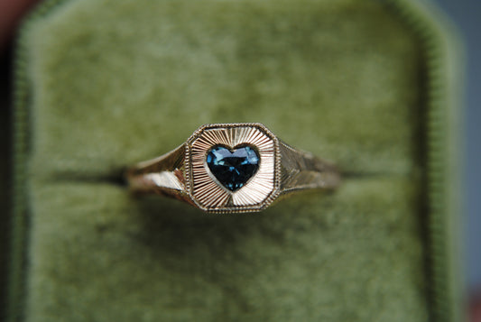 Gold ring with a heart-shaped blue gemstone on a green cushioned background