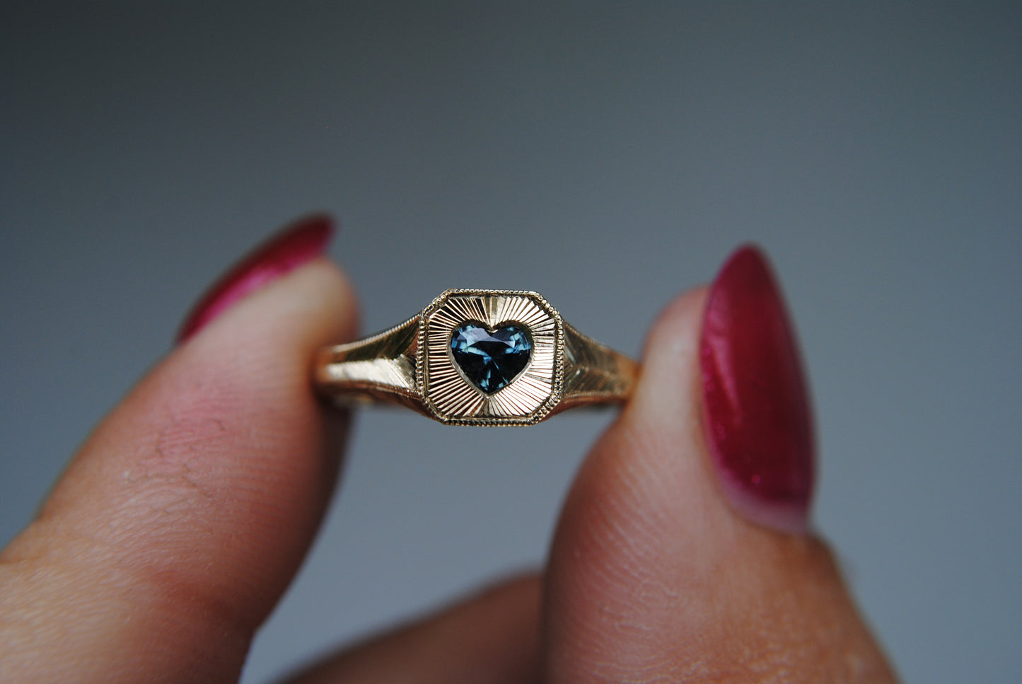 Gold ring with a heart-shaped gem held between fingers against a gray background