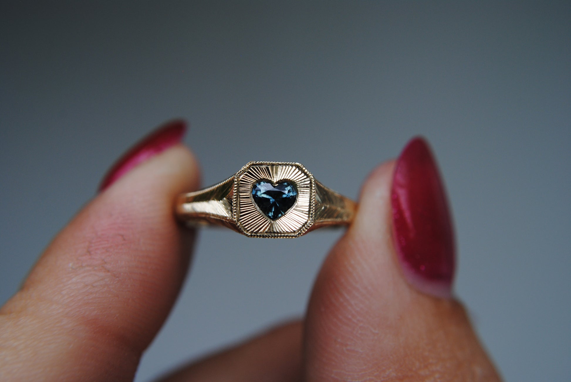 Gold ring with a heart-shaped gem held between fingers against a gray background