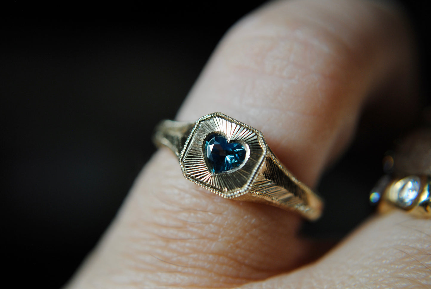 Gold ring with a heart-shaped blue gemstone on a finger against a dark background