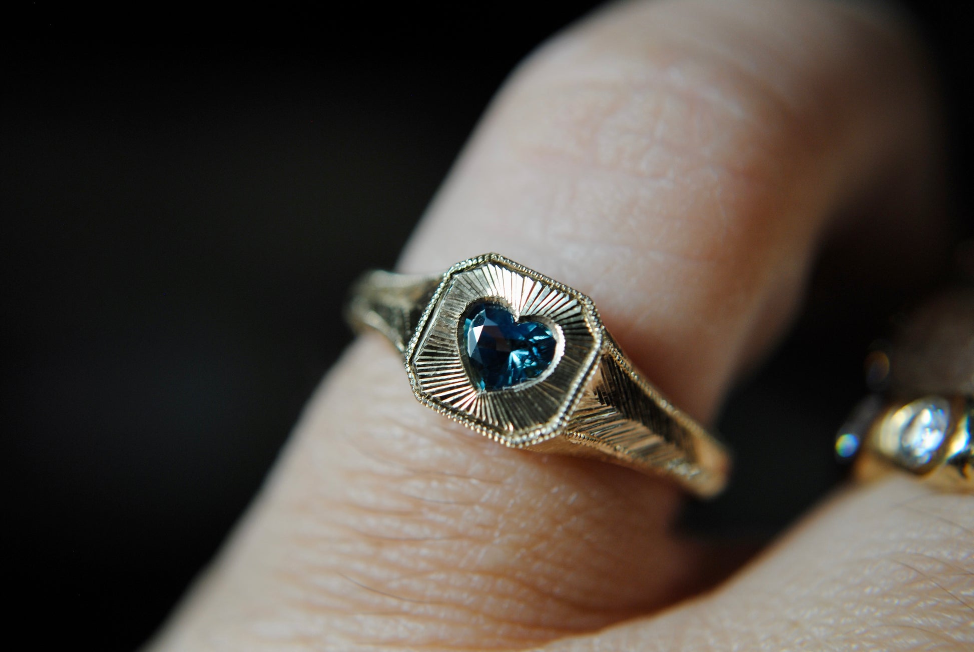 Gold ring with a heart-shaped blue gemstone on a finger against a dark background