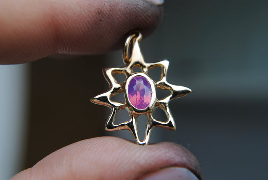 Gold pendant with a purple gemstone held between fingers against a blurred background