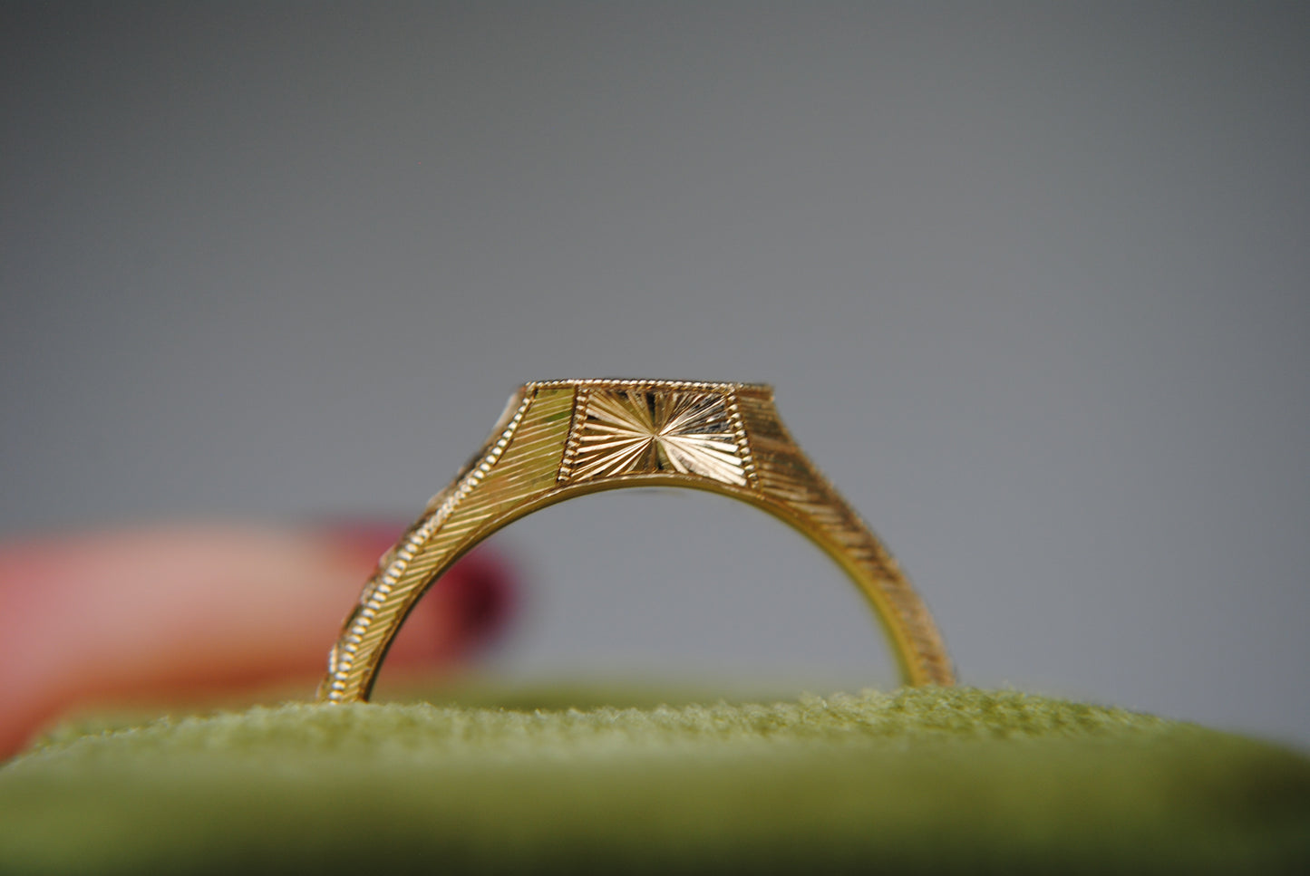 Gold ring with intricate design on a green cushion against a gray background