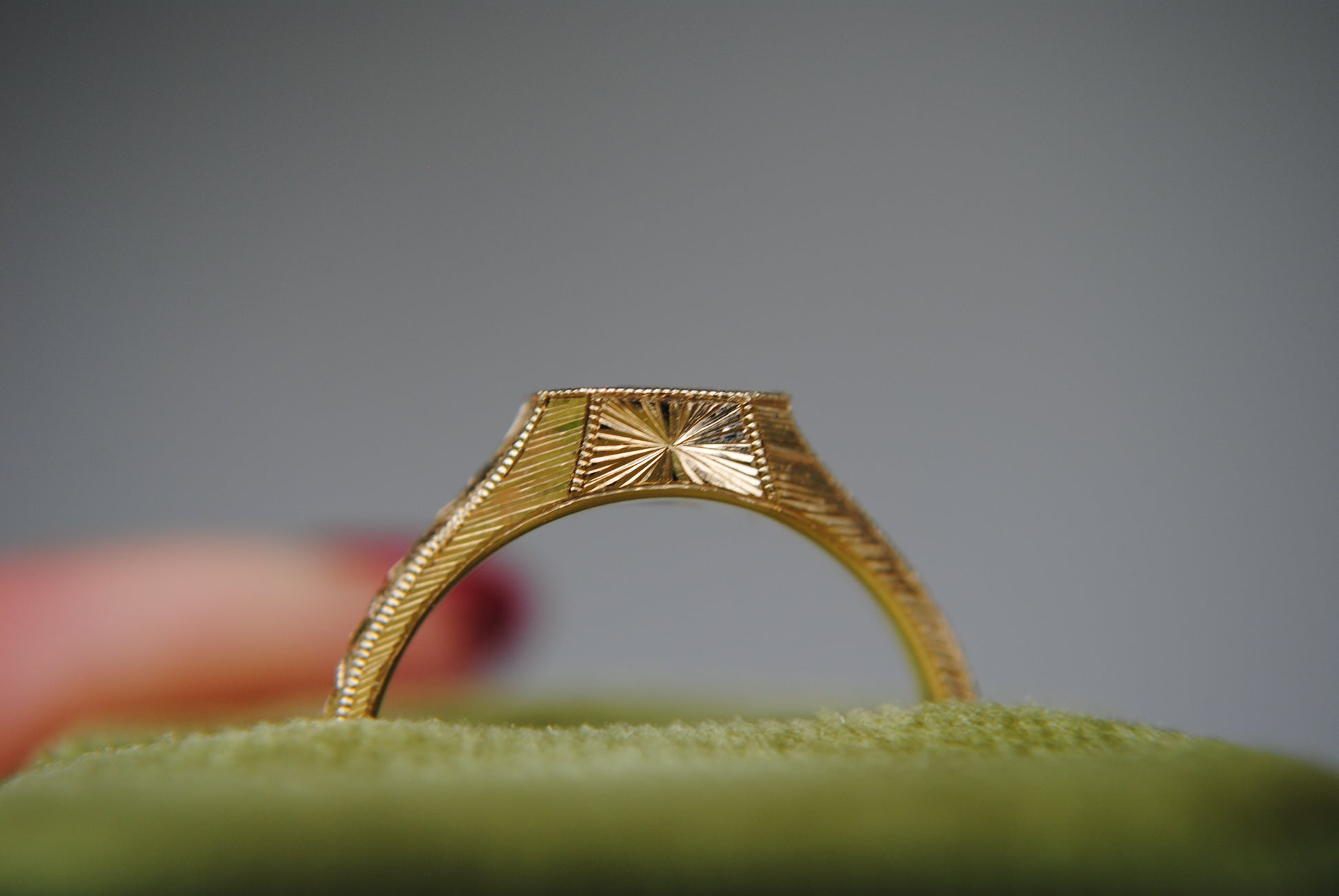 Gold ring with intricate design on a green cushion against a gray background